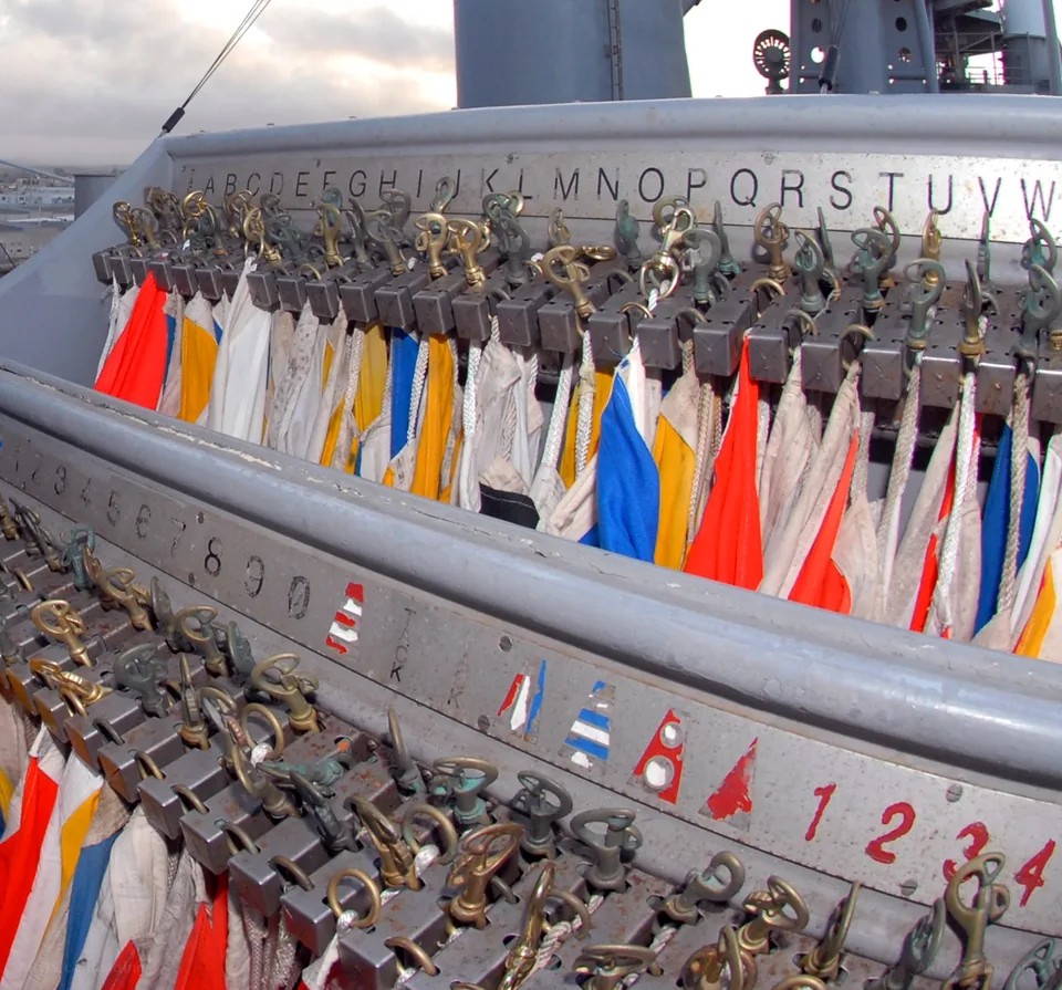 A ship's signal-flag locker with brass clips under painted letters A to W, each nautical signal flag hanging in its own slot