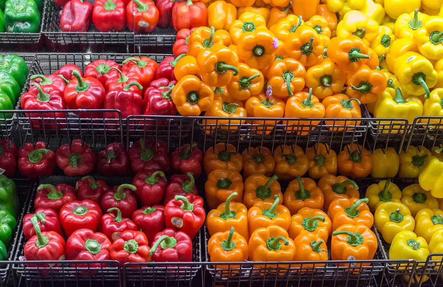 Color-sorted bell peppers in red, orange, yellow and green, arranged in black wire produce baskets in a retail